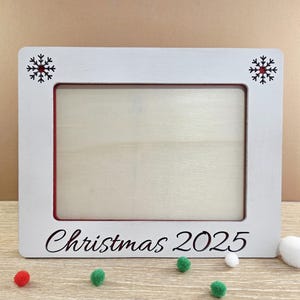 May include: A white wooden picture frame with a red interior border. The frame has black snowflake designs in the corners and the words "Christmas 2025" in script at the bottom. Small decorative pom-poms are scattered around the frame.