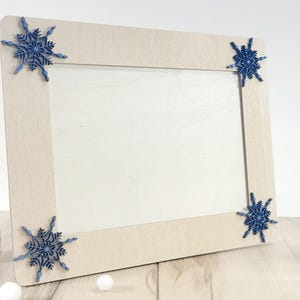 Puede incluir: Un marco de fotos de madera clara con un paspartú blanco y adornos de copo de nieve azul en cada esquina.