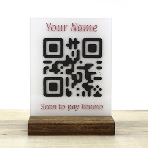 May include: White acrylic sign with a black and white QR code and the text "Your Name" and "Scan to pay Venmo" in pink. The sign is on a dark brown wooden stand.