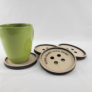 Puede incluir: Cuatro posavasos de madera con borde negro y diseño de botón. El posavasos superior tiene el texto "YOUR NAME GOES HERE" grabado. Una taza verde está sentada en uno de los posavasos.