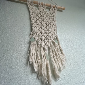 May include: A white macrame wall hanging with a wooden dowel at the top and a fringe at the bottom. The macrame is decorated with small, light green sea glass pieces.