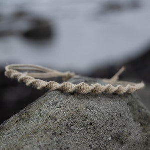 May include: A close-up of a beige rope bracelet on a gray rock. The bracelet is made of a thick, braided rope and has a simple, minimalist design.
