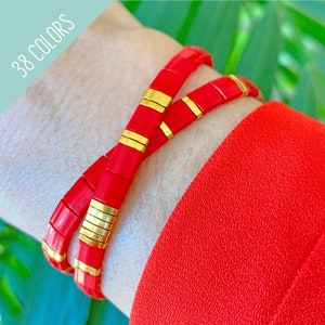 Op de afbeelding: Twee rode en gouden armbanden met een geometrisch ontwerp. De armbanden zijn gestapeld op de pols van een persoon.