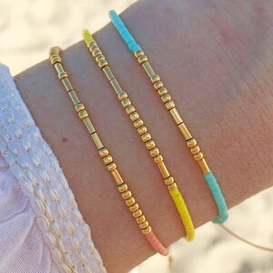 Op de afbeelding: Drie gouden kralen armbanden met draden in verschillende kleuren. De armbanden zijn gestapeld op de pols van een persoon. De draden zijn turquoise, geel en roze.