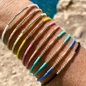 Puede incluir: Una pila de pulseras de cuentas de colores con detalles dorados. Cada pulsera tiene una fina barra dorada y pequeñas cuentas doradas.