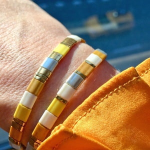 Op de afbeelding: Drie armbanden gestapeld op een pols. De armbanden zijn gemaakt van kleine, vierkante kralen in tinten geel, wit en goud. De kralen zijn gerangschikt in een herhalend patroon.