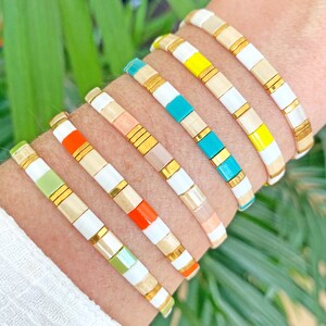 Op de afbeelding: Een set kleurrijke armbanden met witte en gouden kralen. De armbanden zijn gestapeld op de pols van een persoon en hebben verschillende kleuren, waaronder groen, oranje, geel en turquoise.
