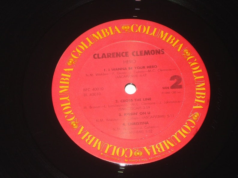 Pu&ograve; includere: Un disco in vinile vintage di Clarence Clemons, intitolato "Hero". L'etichetta rossa presenta il logo Columbia Records e titoli di canzoni come "I Wanna Be Your Hero" e "Kissin' On U". Il disco &egrave; nero con un'etichetta rossa.