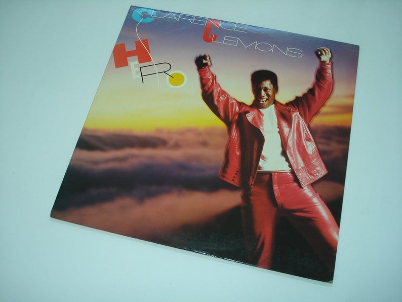 Pu&ograve; includere: Copertina di un album in vinile con Clarence Clemons in abito di pelle rossa con le braccia alzate. L'album si intitola "Hero". Lo sfondo mostra un tramonto sulle nuvole. La copertina dell'album presenta il testo "Clarence Clemons" e "Hero".