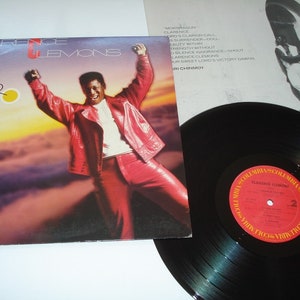 Pu&ograve; includere: Copertina dell'album "Hero" di Clarence Clemons, con un uomo in tuta di pelle rossa con le braccia alzate. Un disco in vinile nero con un'etichetta rossa e oro in primo piano. Il titolo dell'album e il nome dell'artista sono visibili.