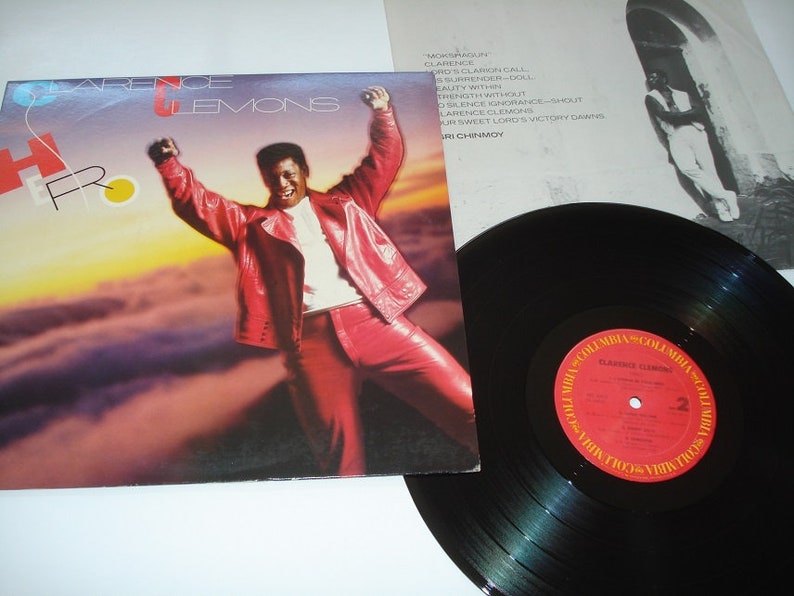 Pu&ograve; includere: Copertina dell'album "Hero" di Clarence Clemons, con un uomo in tuta di pelle rossa con le braccia alzate. Un disco in vinile nero con un'etichetta rossa e oro in primo piano. Il titolo dell'album e il nome dell'artista sono visibili.