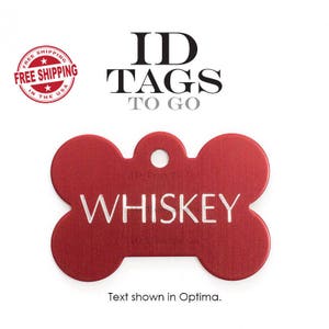 Puede incluir: Una placa de identificaci&oacute;n para perro de hueso rojo con el nombre "WHISKEY" grabado en letras blancas. La placa tiene un agujero para un anillo o una cadena. El texto "ID TAGS TO GO" se muestra encima de la placa.