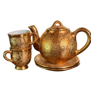 May include: A metallic gold tea set featuring a teapot with a lid, two stacked cups, and two saucers. Each piece has a textured, mottled surface finish. The interior of the cups is white, while the exterior and all other items are gold.