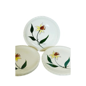 May include: A set of white ceramic plates with a hand-painted floral design. Each plate features a yellow and brown flower with green leaves and stems. The plates have a scalloped edge.