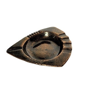 May include: A triangular ashtray with a dark brown and bronze metallic finish. The object features a central circular basin surrounded by a wide, flat rim with three distinct notches for resting items, showcasing a textured, speckled surface.