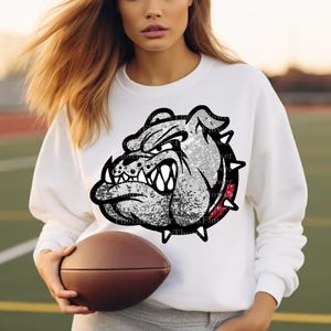 Peut inclure: Sweat-shirt blanc avec un graphique de bouledogue en paillettes argentées. Le bouledogue a un collier rouge et une expression féroce. Le sweat-shirt est porté par une personne tenant un ballon de football brun.