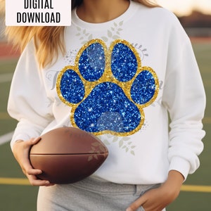 Peut inclure: Un sweat-shirt blanc avec un motif d'empreinte de patte en paillettes bleues et dorées. L'empreinte de patte est bordée d'or et a un centre en paillettes dorées. Le sweat-shirt est porté par une personne tenant un ballon de football brun.