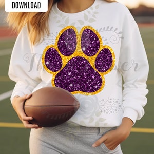 Peut inclure: Sweat-shirt blanc avec un motif d'empreinte de patte en paillettes violettes et dorées. Le sweat-shirt est porté par une personne tenant un ballon de football brun.