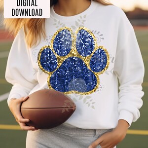 Puede incluir: Una sudadera blanca con un diseño de huella de pata de purpurina azul y dorada. La huella de pata está bordeada de dorado y tiene un centro de purpurina dorada. La sudadera es una descarga digital.