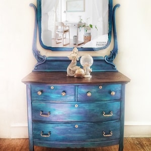 May include: A vintage dresser with a curved front and four drawers. The dresser is painted in a teal blue and purple ombre finish with gold hardware. The dresser has a mirror with an ornate frame and a mermaid figurine on top.