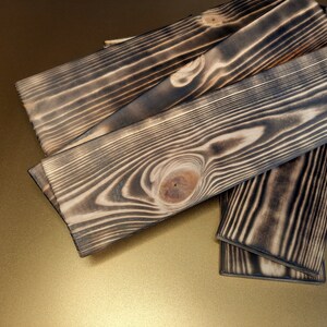 May include: Four pieces of wood with a dark, charred finish. The wood has a natural grain pattern and a knot in the center. The wood is stacked on top of each other.