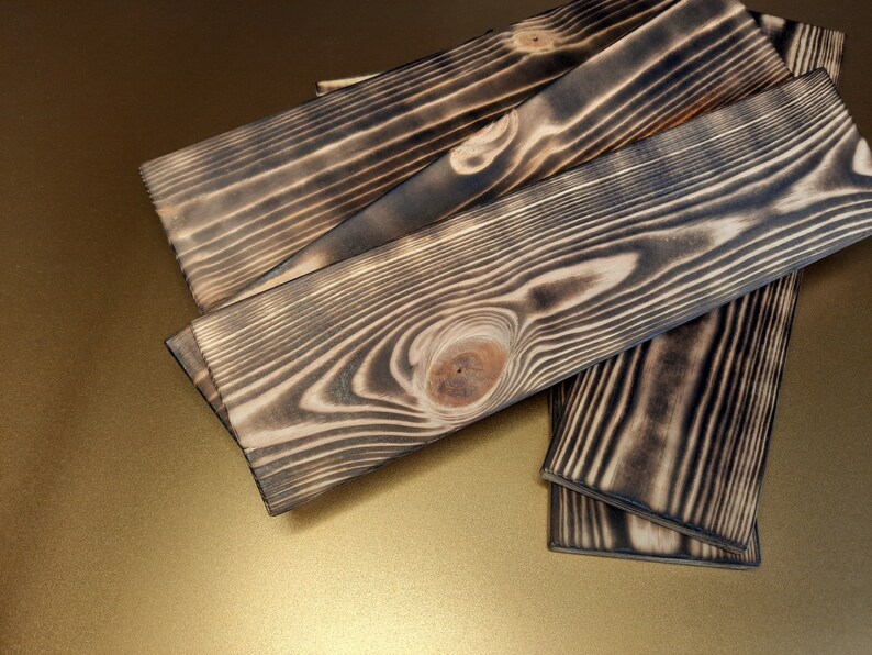 May include: Four pieces of wood with a dark, charred finish. The wood has a natural grain pattern and a knot in the center. The wood is stacked on top of each other.