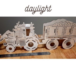 May include: A wooden model of a tractor with a trailer. The tractor has a scoop on the front and the trailer has a bed with a textured surface. The model is painted in a natural wood color.