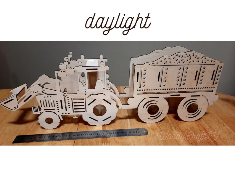 May include: A wooden model of a tractor with a trailer. The tractor has a scoop on the front and the trailer has a bed with a textured surface. The model is painted in a natural wood color.