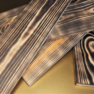 May include: Four pieces of dark brown wood planks with a charred finish. The wood grain is visible and the planks are stacked on top of each other.