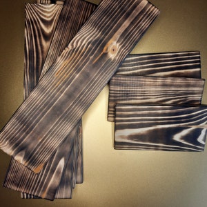 May include: A set of six wooden planks with a dark brown, charred finish. The planks are arranged in a fan shape, with the largest plank at the top and the smallest plank at the bottom.