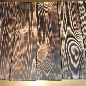 May include: Four wooden planks with a dark brown, burnt wood finish. The planks have a natural wood grain pattern and are arranged side by side.