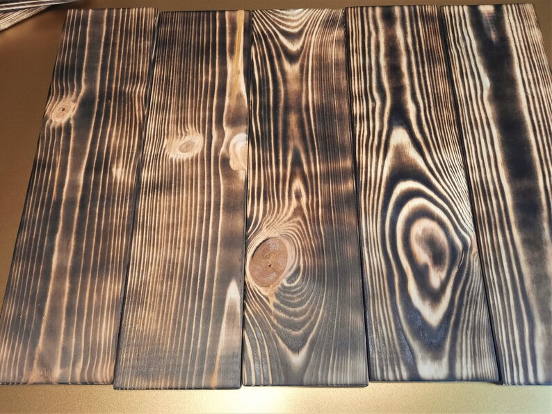May include: Four wooden planks with a dark brown, burnt wood finish. The planks have a natural wood grain pattern and are arranged side by side.