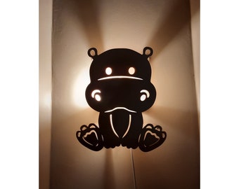 May include: A black silhouette of a hippopotamus with a light shining through the cut-out eyes and mouth. The hippo is mounted on a wall and is a nightlight.