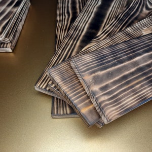 May include: Close-up of several wooden planks with a dark, charred finish. The planks have a natural wood grain pattern and are arranged in a fan shape.