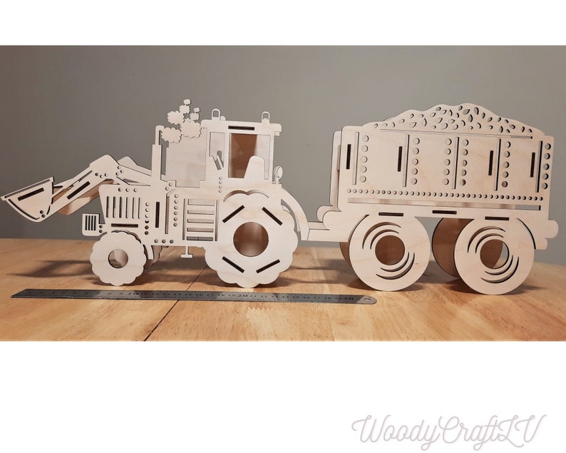 May include: A wooden model of a tractor with a trailer. The tractor has a scoop on the front and the trailer has a bed filled with dirt. The model is made of unfinished wood.
