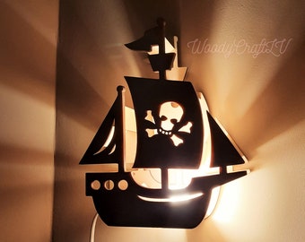 May include: A black wooden pirate ship-shaped wall lamp with a skull and crossbones design on the sail. The lamp is illuminated from within, casting a warm glow. The lamp is mounted on a light-colored wall.