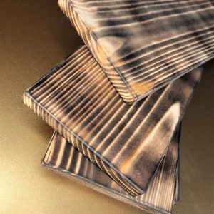 May include: Three wooden boards with a dark brown, charred finish. The boards are stacked on top of each other, with the top board slightly offset from the bottom two.