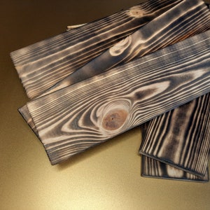 May include: Four pieces of wood with a dark, charred finish. The wood has a natural grain pattern and a knot in the center. The wood is stacked on top of each other.