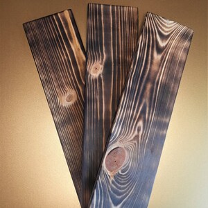 May include: Three dark brown wooden planks with a wood grain pattern and knots. The planks have a charred, burnt look.