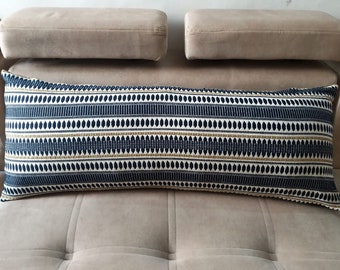 May include: A long, rectangular throw pillow with a geometric pattern in shades of navy blue, white, and gold. The pillow is on a light brown sofa.