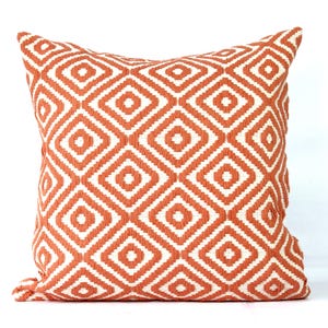 May include: Square throw pillow with a geometric diamond pattern in orange and white. The pillow has a textured appearance and is likely made of fabric. The design features repeating diamond shapes.