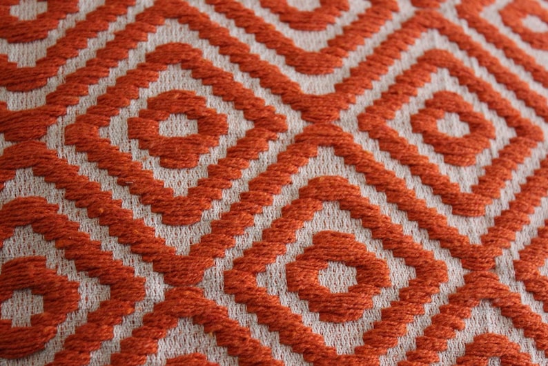 K&ouml;nnte beinhalten: Orangefarbener und wei&szlig;er gewebter Stoff mit einem geometrischen Muster. Das Muster besteht aus kleinen Quadraten und Kreisen.