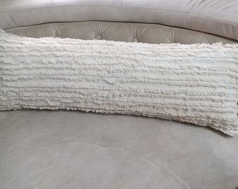 May include: A white, long, rectangular pillow with a fringed texture. The pillow is lying on a light gray couch.