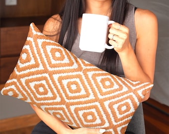 Può includere: Una donna tiene in mano una tazza di ceramica bianca e un cuscino con motivo marrone e bianco. Il cuscino ha un design geometrico con quadrati e rombi.