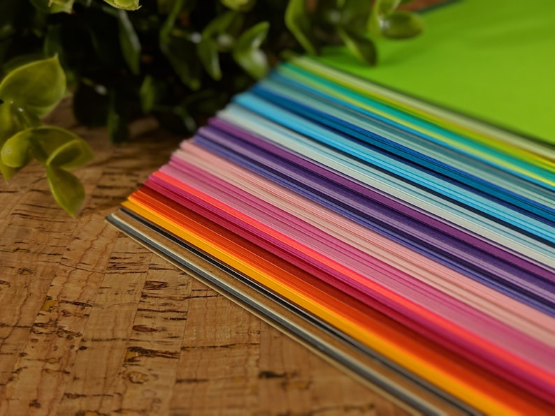 May include: A vibrant array of colorful paper sheets, fanned out to display a spectrum of hues. The colors range from warm reds and oranges to cool blues and greens, creating a visually appealing gradient. The paper is neatly stacked, suggesting a craft or stationery product.