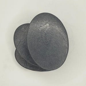 May include: A stack of three dark gray, oval-shaped coasters with a textured, matte finish. The coasters are layered slightly offset from one another, creating a tiered appearance. Each piece has a consistent dark charcoal color and a smooth, rounded edge.