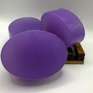 Könnte beinhalten: Drei ovale, violette Seifen werden gezeigt. Die Seifen haben eine glatte, matte Oberfläche und sind auf einer kleinen Seifenschale aus Holz angeordnet. Die Seifen haben einen leuchtenden Lilaton, und die Holzschale bietet einen natürlichen Kontrast.