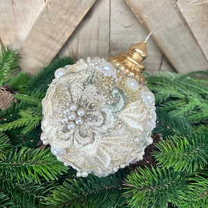 Pu&ograve; includere: Una decorazione natalizia sferica in pizzo color crema con ricami floreali, arricchita da piccole perle bianche e dettagli glitterati dorati. L'ornamento &egrave; sormontato da un tappo in metallo dorato e da un filo di sospensione trasparente.