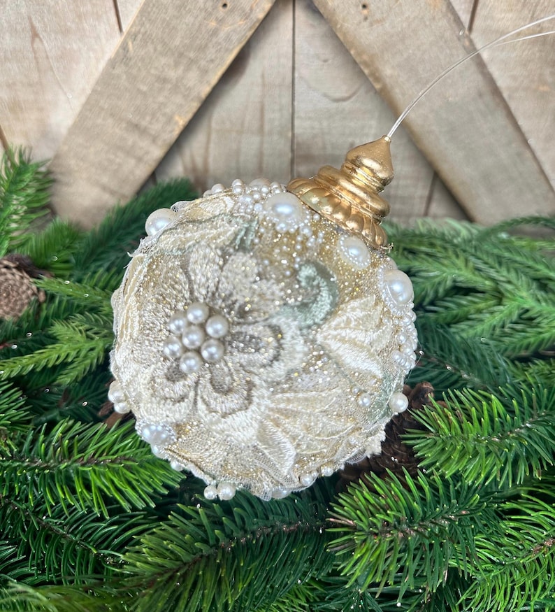 Pu&ograve; includere: Una decorazione natalizia sferica in pizzo color crema con ricami floreali, arricchita da piccole perle bianche e dettagli glitterati dorati. L'ornamento &egrave; sormontato da un tappo in metallo dorato e da un filo di sospensione trasparente.