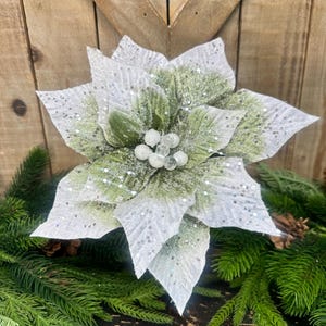 May include: A decorative poinsettia flower featuring white petals with silver glitter accents, layered over sage green leaves. The center of the flower is adorned with a cluster of small, round, translucent white beads, creating a textured appearance.